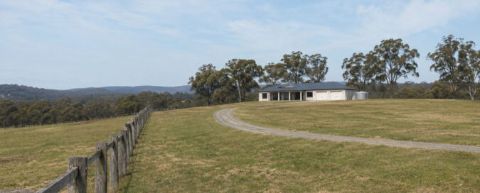 Wide view across a rural residential block in the Hawkesbury region near Sydney, a single-storey homestead with a triple garage set back on the rise, paddocks in front and a wooded ridge behind under a hazy blue sky
