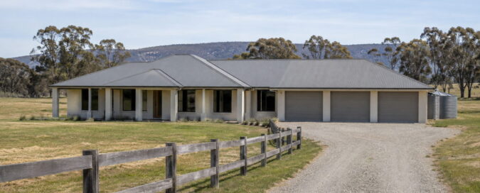 Completed single-storey acreage home in the Hawkesbury with a hipped Colorbond roof, wide verandah and three-car garage, viewed from the gravel driveway on a clear afternoon with paddock and gum trees behind