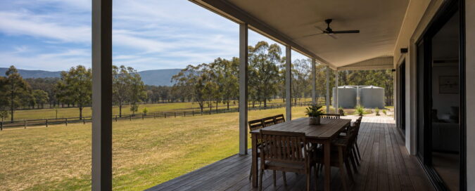 Deep covered verandah of a single-storey acreage home looking out over a mown paddock to gum trees, with an outdoor dining table under the roof and rainwater tanks visible at the side of the house